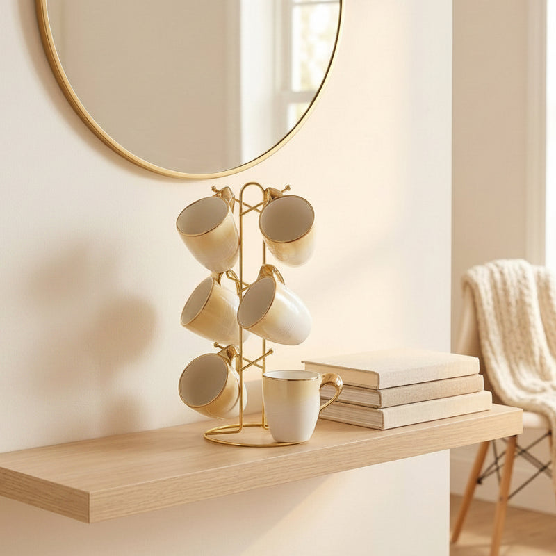 A gold-tone mug rack holding six cream ceramic mugs, styled on a light wood floating shelf next to a potted olive tree and books, with a large round mirror and a chair in the background.